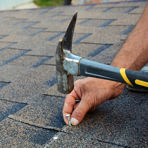 Worker hands installing bitumen roof shingles. Worker Hammer in Nails on the Roof. Roofer is hammering a Nail in the Roof Shingles. Unfinished roof.