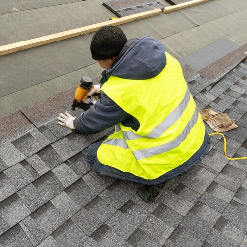 Qualified workman in uniform work wear using air or pneumatic nail gun and installing asphalt shingle roofing